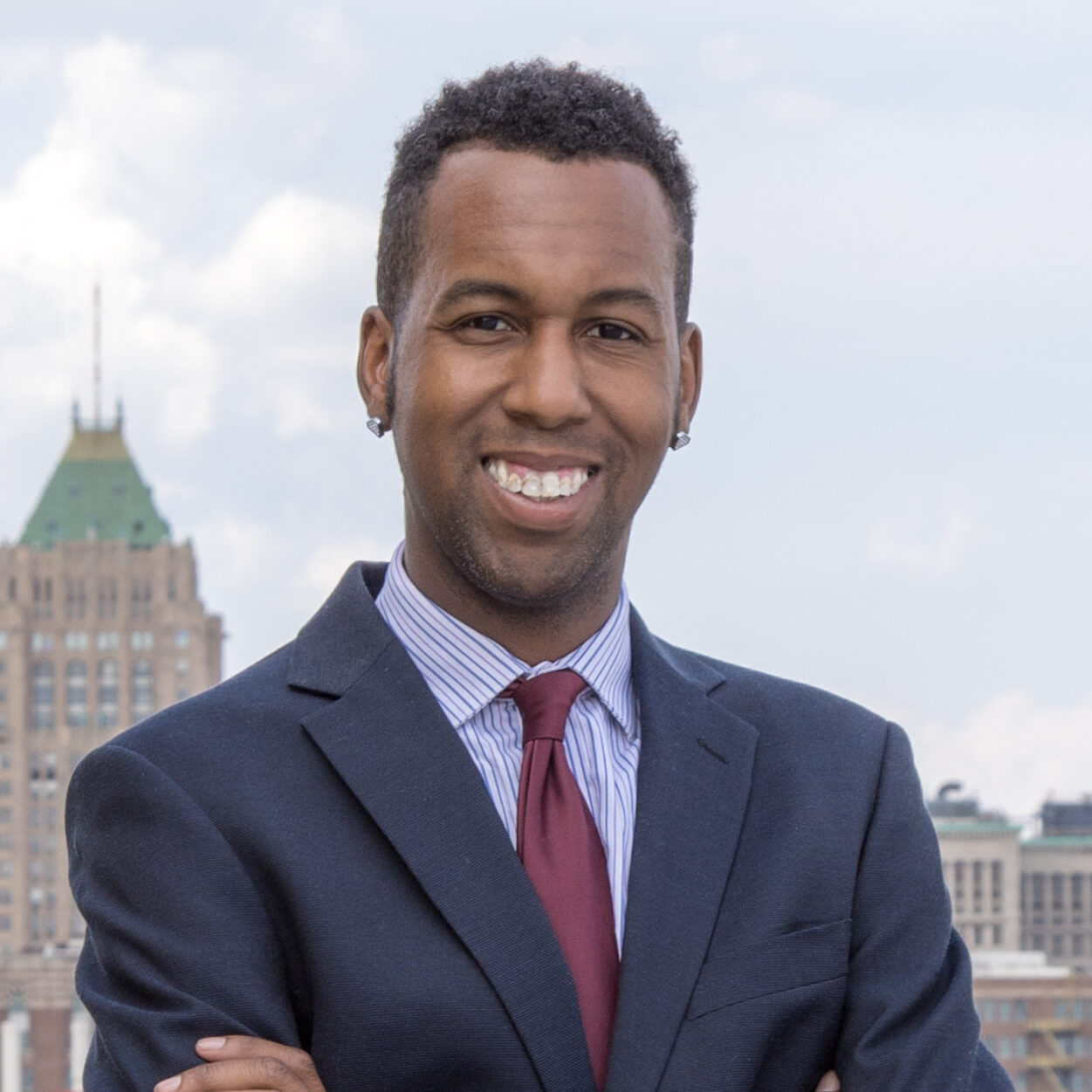 Michigan LCV Voting Rights Coordinator Roland Leggett smiling at the camera on a roof in Midtown, Detroit. He is a queer man of color wearing a suit.